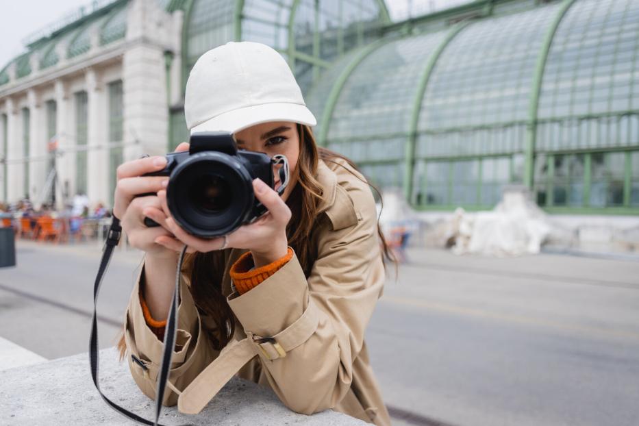 Eine Frau mit beiger Jacke und weißer Kappe hält eine Kamera und fotografiert vor dem Palmenhaus im Burggarten in Wien.