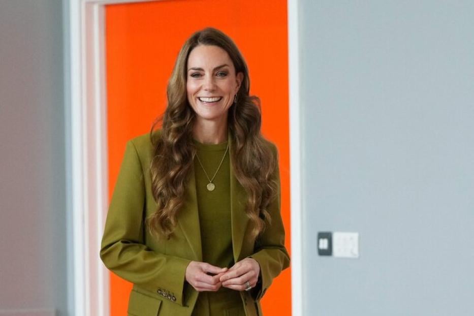 Prinzessin Kate trägt einen olivgrünen Hosenanzug und steht lächelnd in einem Raum mit orangefarbener Tür.