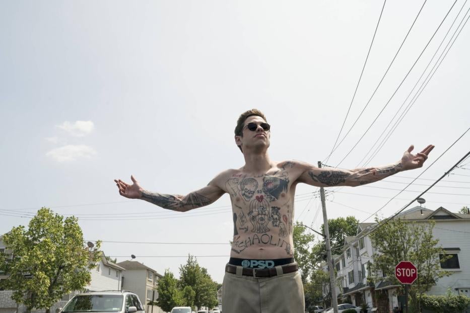 Szene aus dem Film The King of Staten Island (2020): Pete Davidson steht mit nacktem Oberkörper, der mit Tattoos bedeckt ist, und ausgebreiteten Armen und Sonnenbrille auf einer Straße in einem Wohngebiet.