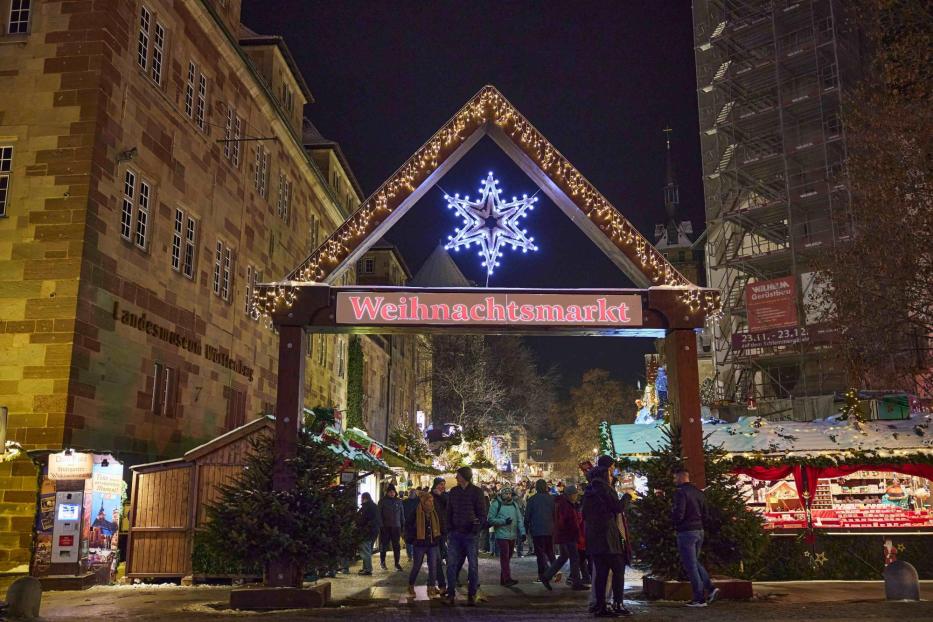 Eingang zum Weihnachtsmarkt mit beleuchtetem Schneeflockensymbol und Schriftzug 'Weihnachtsmarkt', Menschen und Stände bei Nacht.