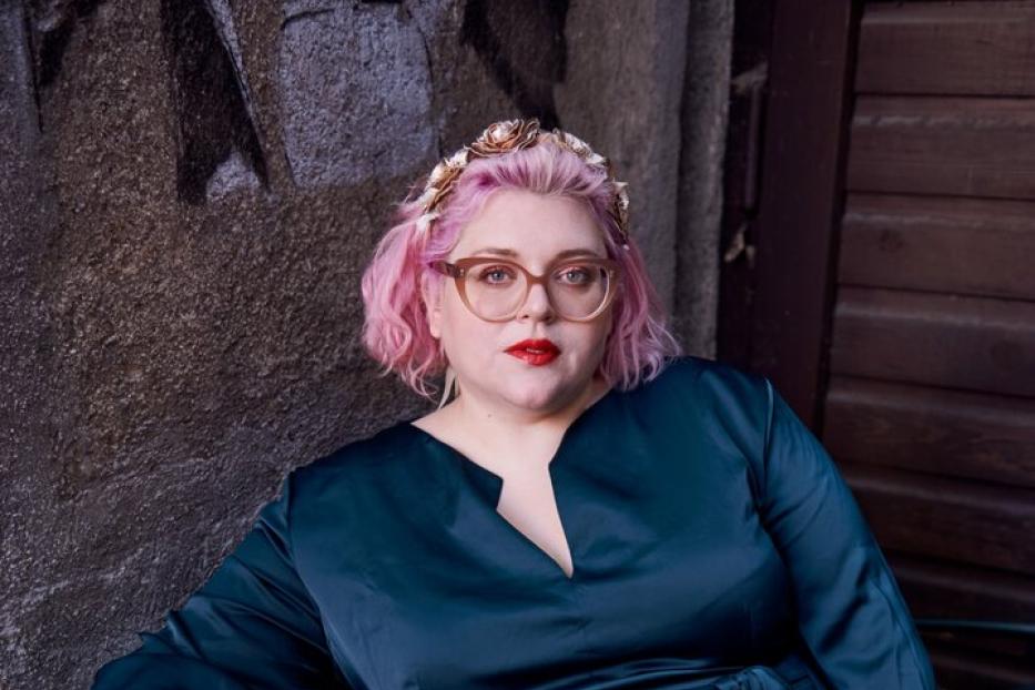 Bobby Herrmann-Thurner sitzt auf einer Bank, trägt einen dunkelblauen Jumpsuit und rosa Haare.