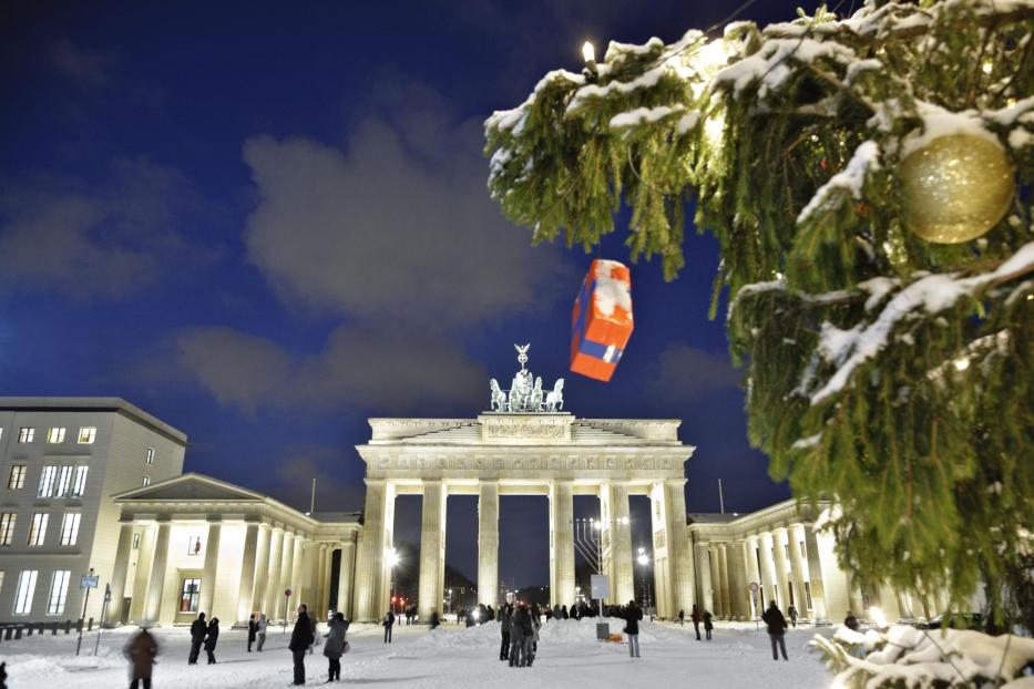 Brandenburger Tor bei Nacht mit Schnee, davor Menschen und ein Weihnachtsbaum mit goldener Kugel im Vordergrund.