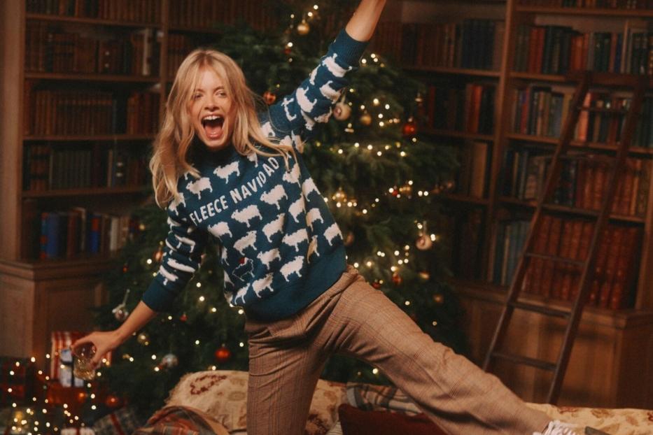 Eine blonde Frau im Weihnachtspullover mit Schafen jubelt vor einem Weihnachtsbaum und Bücherregalen.