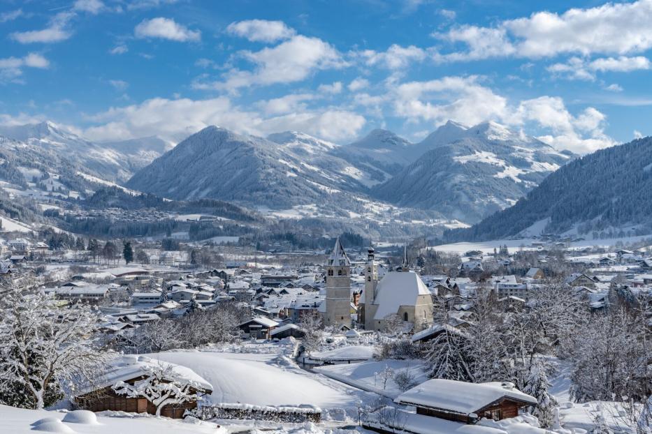 Blick von oben ins Tal auf Kitzbühel: Zwei Kirchen ragen heraus, dahinten erheben sich die Berge. Die Landschaft ist verschneit.