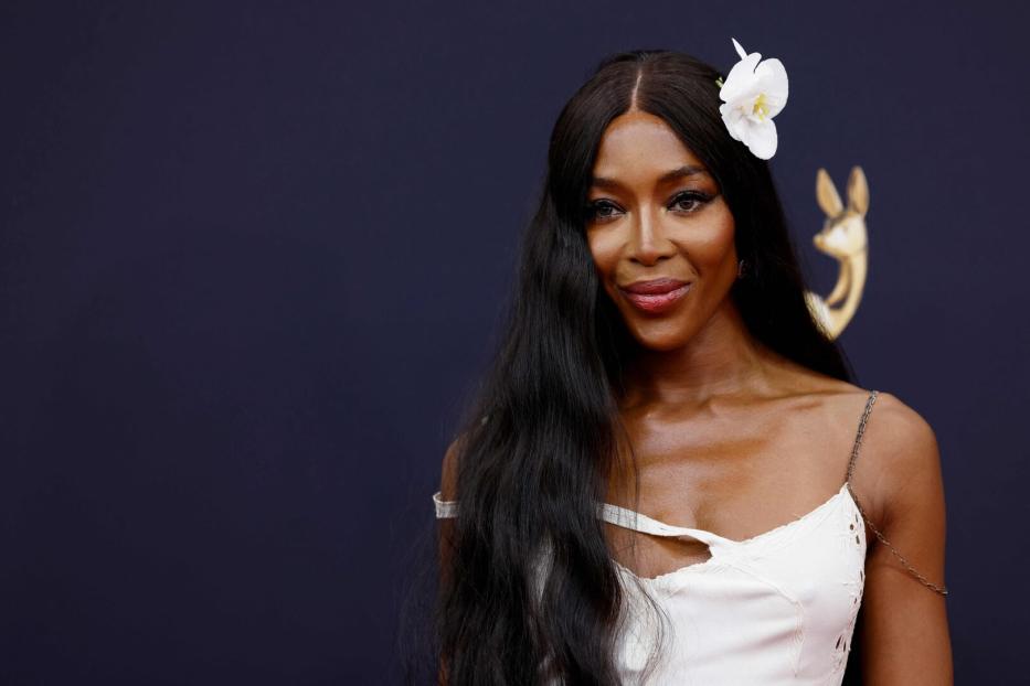 Naomi Campbell mit weißer Blüte im Haar und weißem Kleid vor dunklem Hintergrund.