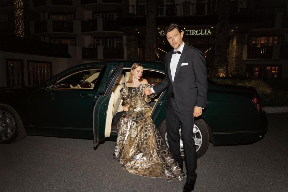 Ein Mann und eine Frau, die aus einem Auto steigen. Er trägt einen Smoking, sie ein goldenes Kleid. 