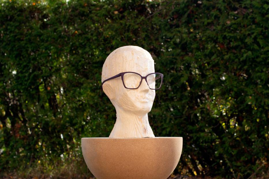 Holzskulptur eines Kopfes mit Brille, stehend in einer großen, runden Schale vor grünem Laub.