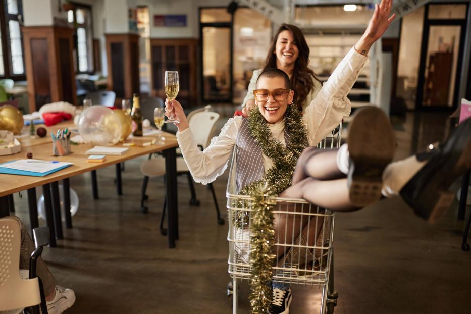 Zwei lachende Personen feiern ausgelassen, eine sitzt mit Sektglas und Girlande in einem Einkaufswagen, die andere schiebt sie.