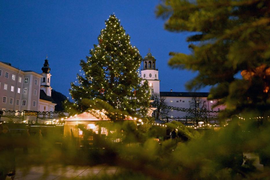 Ein Weihnachtsbaum mit Lichtern und Sternen steht auf einem Platz in Salzburg vor Gebäuden.