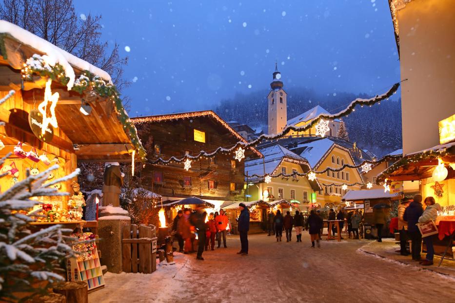 Weihnachtsmarkt mit beleuchteten Holzständen, schneebedecktem Boden und Kirche im Hintergrund bei Abenddämmerung.