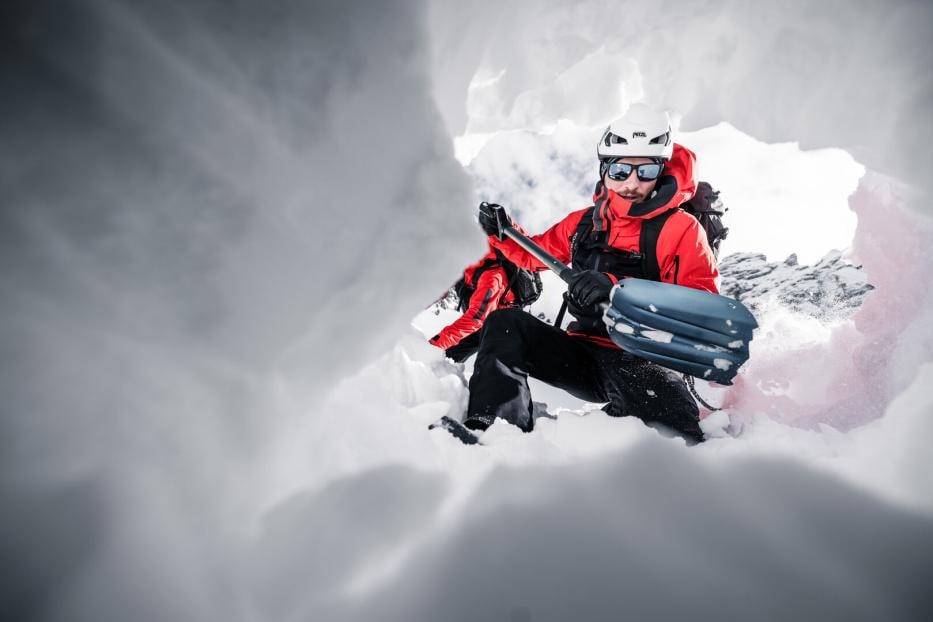 Zwei Personen in roter Winterkleidung mit Skiausrüstung in einer Schneehöhle, umgeben von Eiswänden.