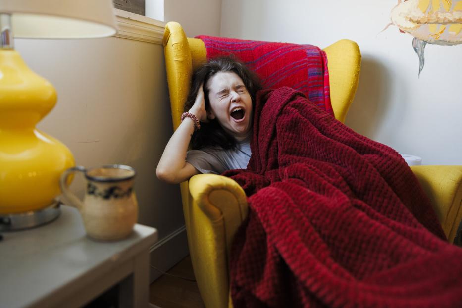Eine Frau gähnt müde in einem gelben Sessel unter einer roten Decke.