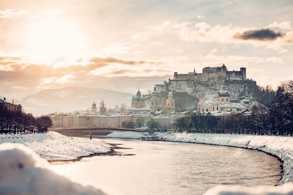 Verschneite Stadt mit Fluss, historischen Gebäuden und einer Burg auf einem Hügel im Sonnenlicht.