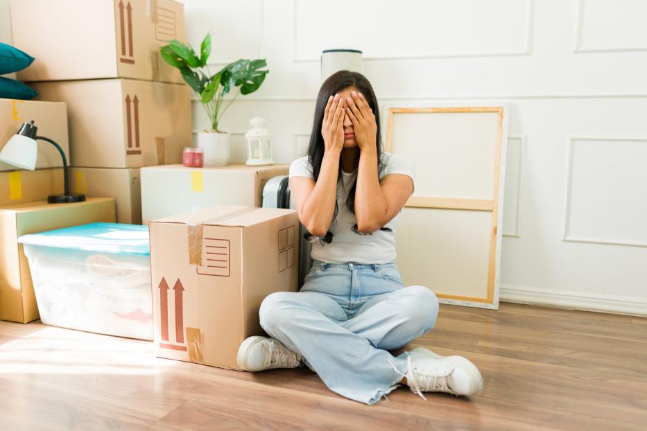 Eine Frau sitzt auf dem Boden zwischen Umzugskartons und bedeckt ihr Gesicht mit den Händen.