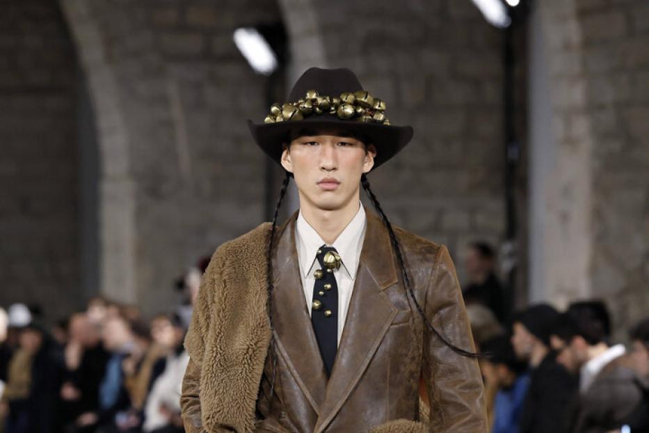 Ein Model mit Hut und Zöpfen präsentiert braune Lederkleidung auf einem Laufsteg.