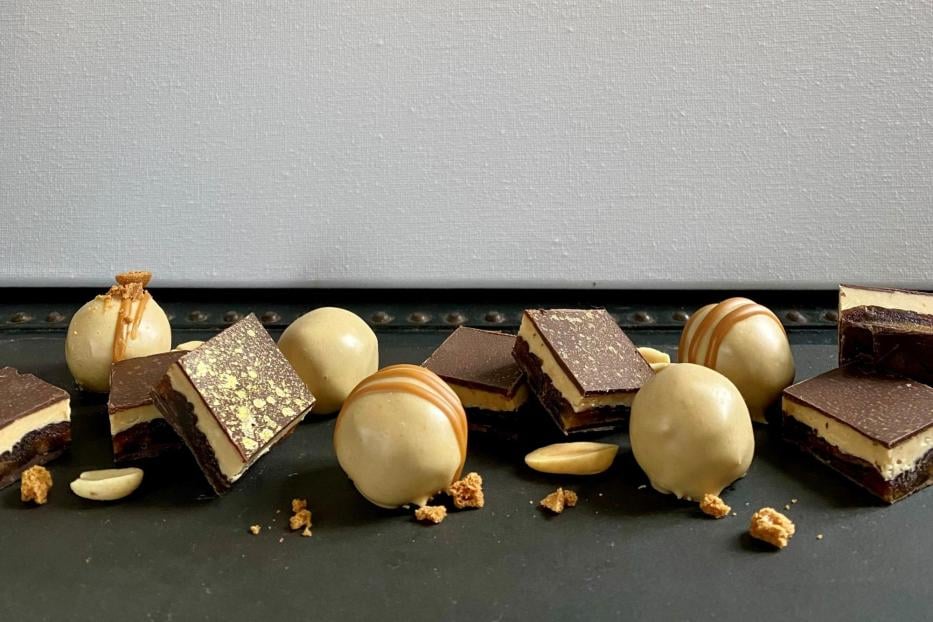 Mehrere Pralinen und Schokoladenstücke mit goldenen Verzierungen und Erdnüssen liegen auf einer dunklen Oberfläche.