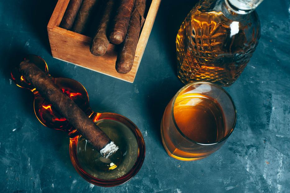 Eine brennende Zigarre liegt in einem Aschenbecher neben einem Glas und einer Karaffe mit braunem Getränk sowie einer Zigarrenkiste.