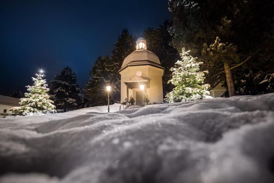Verschneite kleine Kapelle bei Nacht, umgeben von beleuchteten Tannenbäumen und Laternen im Schnee.