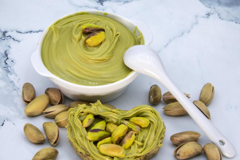 Herzförmiges Brotstück mit Pistaziencreme und Pistazien, daneben eine Schale mit Creme und ein Löffel, umgeben von Pistazien.