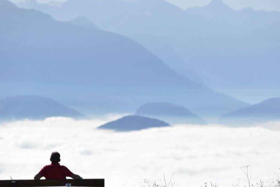 Eine Person sitzt auf einer Bank und blickt auf eine weite Berglandschaft im Nebel.