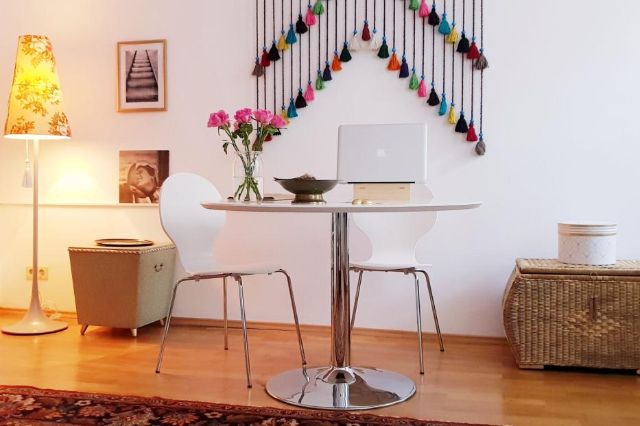 Runder Tisch mit zwei weißen Stühlen, darauf ein Laptop und Blumen, daneben bunte Wanddeko und Bücherregal.