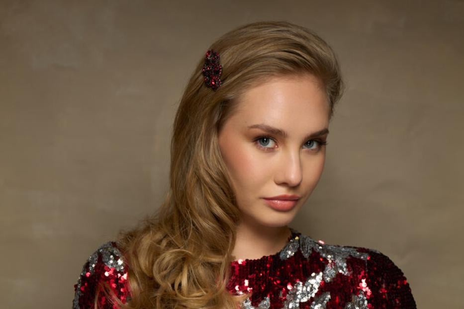 Eine Frau mit blonden Locken trägt ein glitzerndes, rot-silbernes Kleid und eine Haarspange.