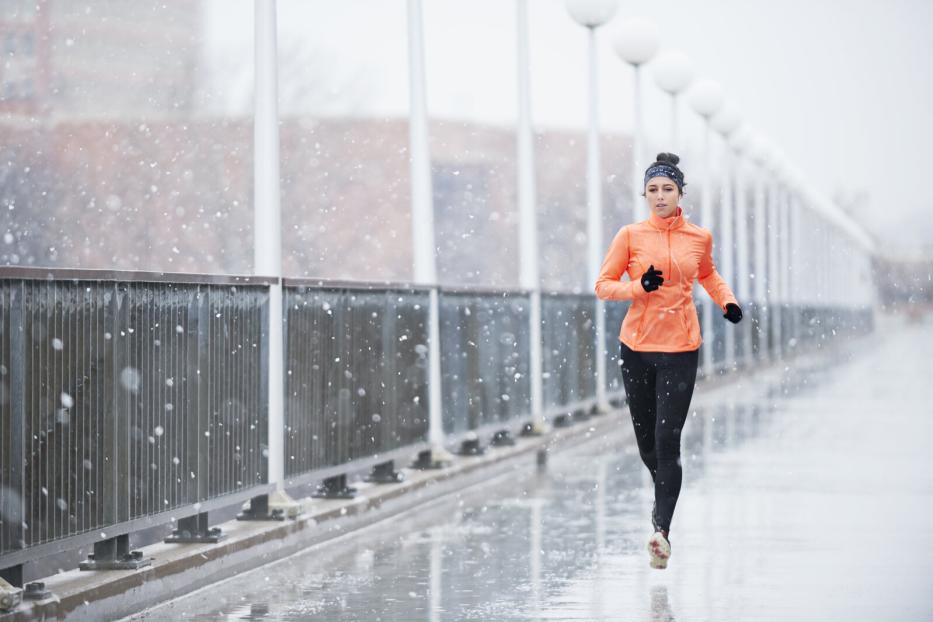 Eine Frau joggt bei Schneefall auf einer Brücke und trägt eine orange Jacke sowie schwarze Leggings.