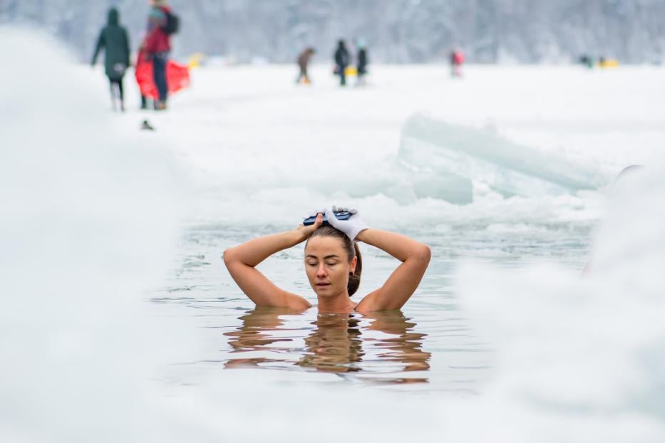 Eine Frau badet in einem eisigen See, umgeben von Schnee, und hält ihre Hände mit Handschuhen auf dem Kopf.