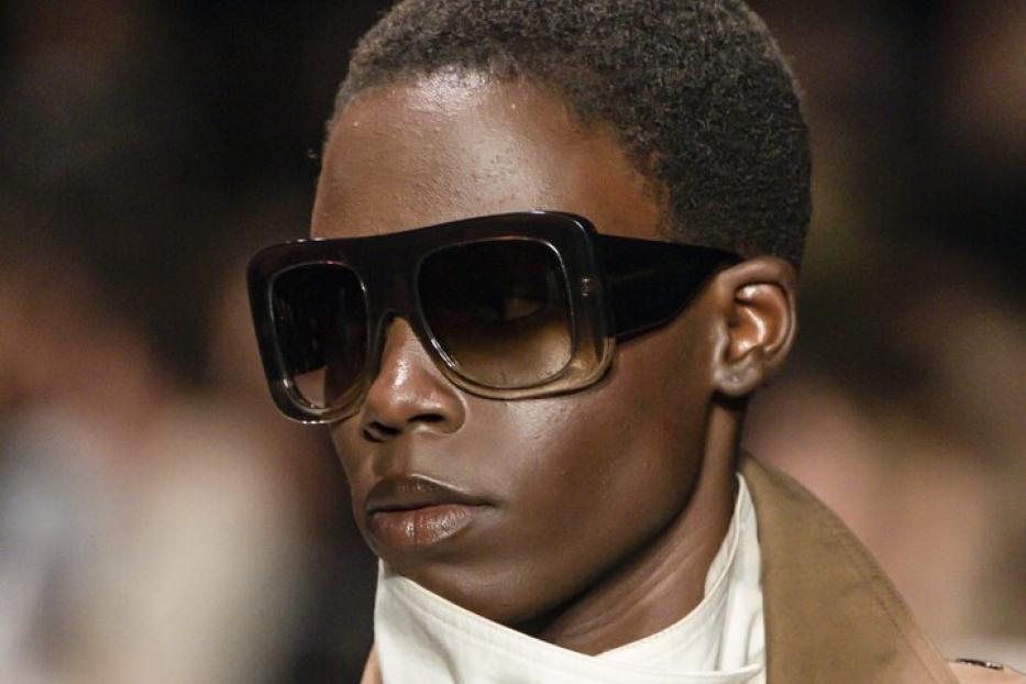 Person mit großer, dunkler Sonnenbrille, kurzem Haar, beigem Mantel und weißem, hochgeschlossenem Oberteil bei der Pariser Fashion Week