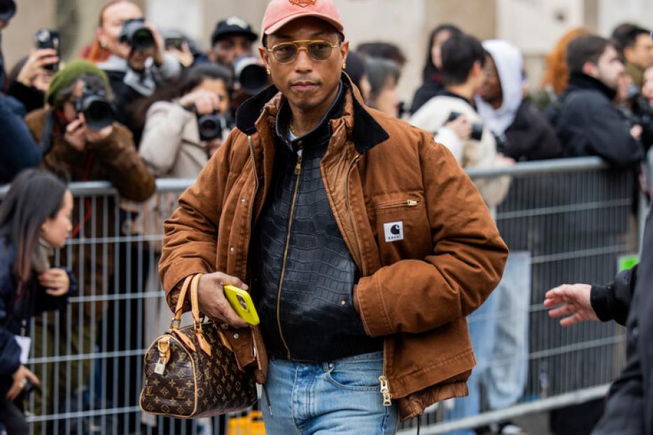 Pharrell Williams trägt eine braune Jacke, Schlaghosen und eine Louis Vuitton Tasche.