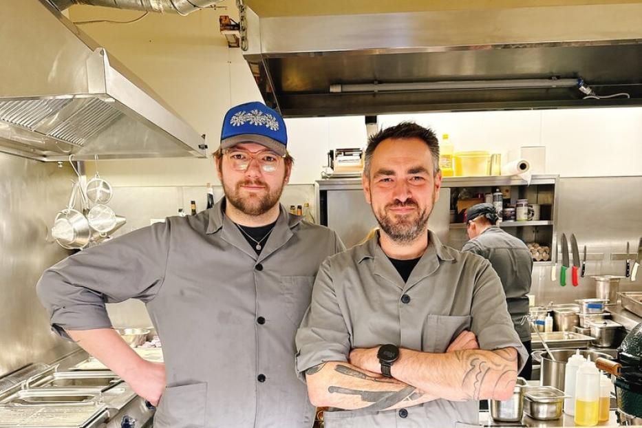 Zwei Köche in grauen Jacken stehen nebeneinander in einer professionellen Restaurantküche.