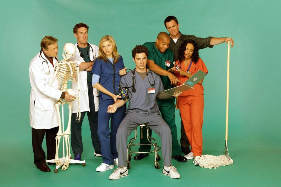 Sieben Personen in Krankenhauskleidung stehen zusammen, eine Person sitzt, ein Skelett und ein Wischmopp sind zu sehen.