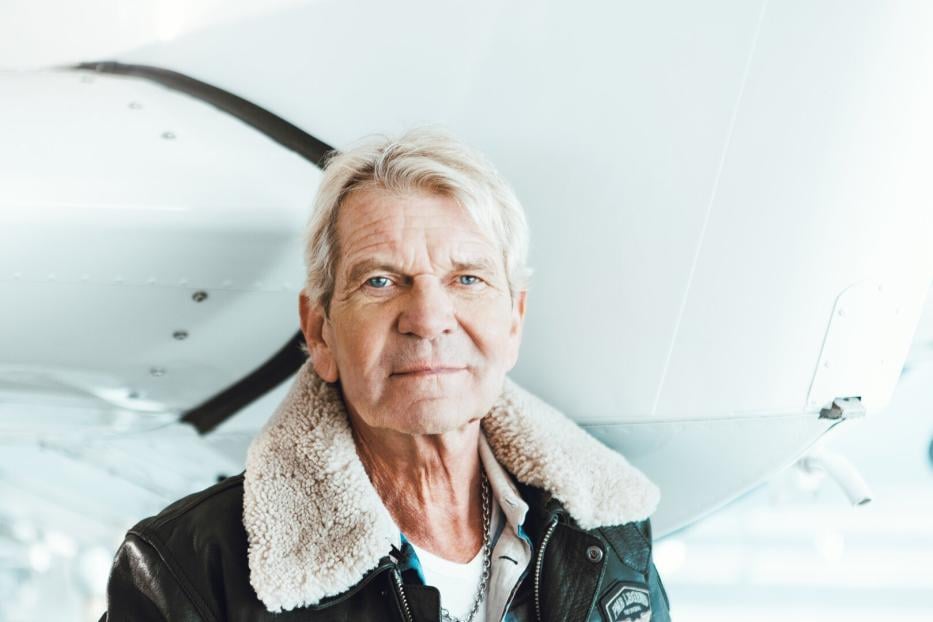 Ein älterer Mann mit grauen Haaren und Fliegerjacke steht vor einem weißen Flugzeug.