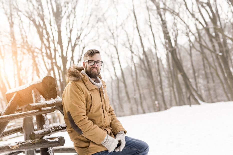 Ein Mann mit Brille und Winterjacke sitzt auf einem Holzzaun im verschneiten Wald.
