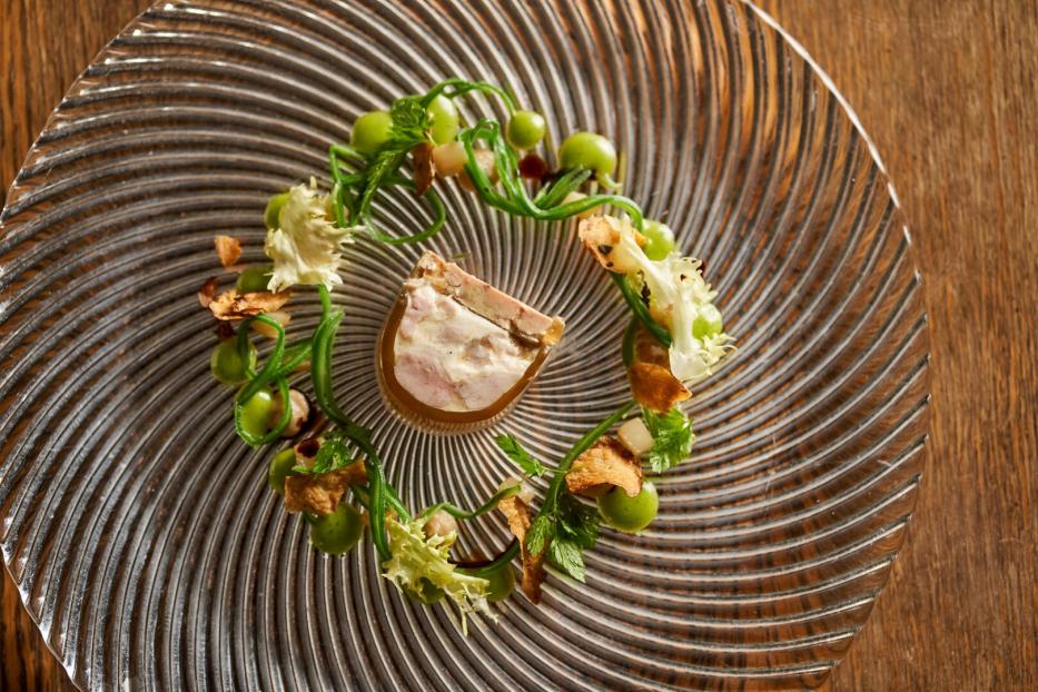 Ein kunstvoll angerichtetes Gourmetgericht mit Terrine, grüner Creme, Kräutern und Salat auf einem gläsernen Teller.