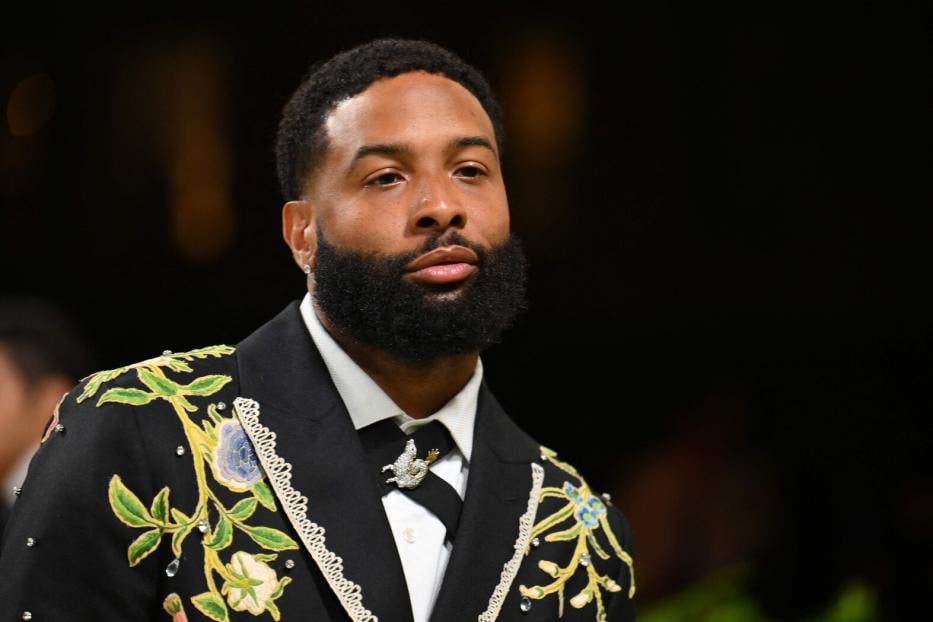 Ein Mann mit Vollbart trägt einen schwarzen Anzug mit floralen Stickereien und Schmuckdetails.