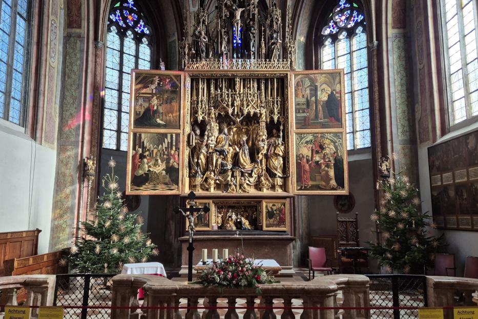 Ein Altar mit goldenem Altaraufsatz und Gemälden steht in einer Kirche mit Weihnachtsbäumen.