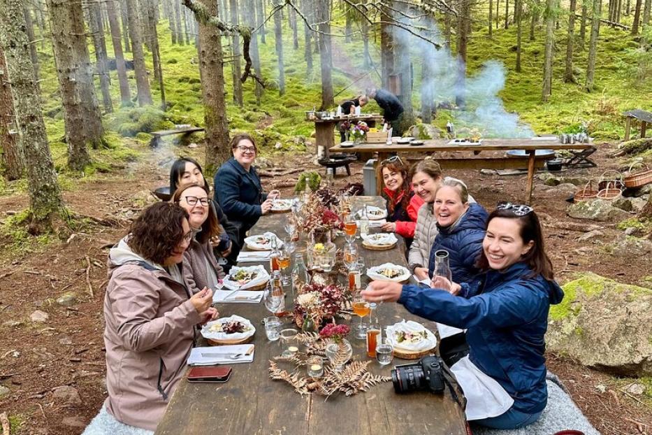 Eine Gruppe von Menschen sitzt an einem rustikalen Holztisch im schwedischen Wald und genießt gemeinsam ein Essen.