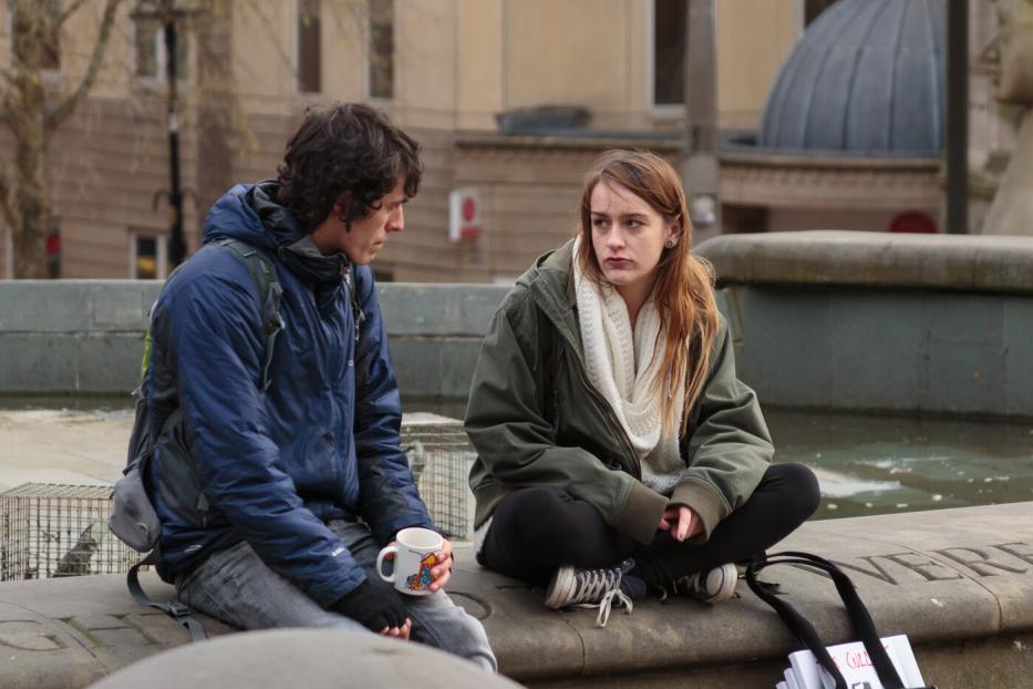 Zwei junge Menschen sitzen auf einer steinernen Umrandung, einer hält eine Tasse, beide wirken nachdenklich.