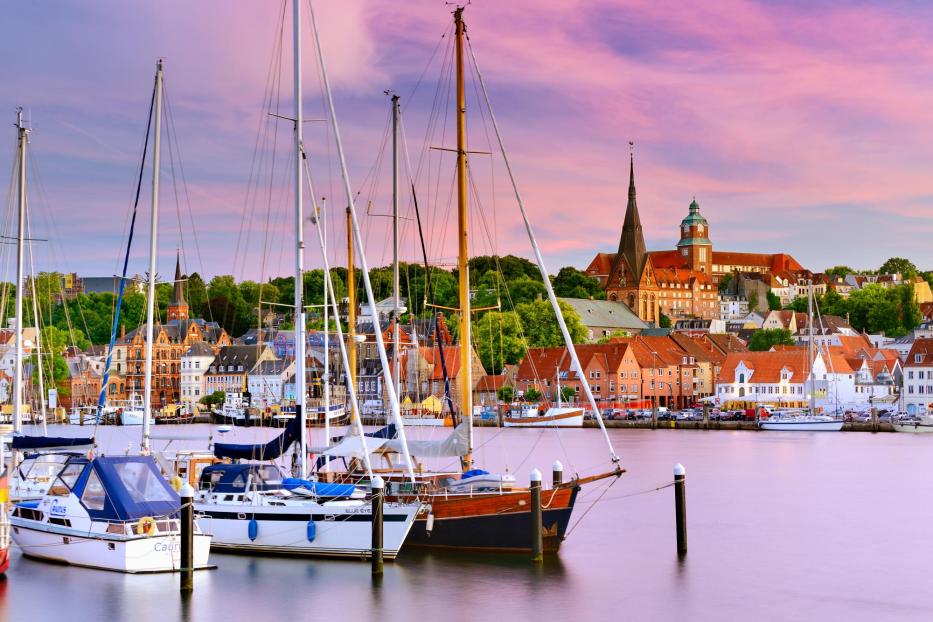 Segelboote liegen im Hafen von Flensburg vor der Kulisse der Altstadt mit historischen Gebäuden und der St.-Nikolai-Kirche bei Sonnenuntergang.