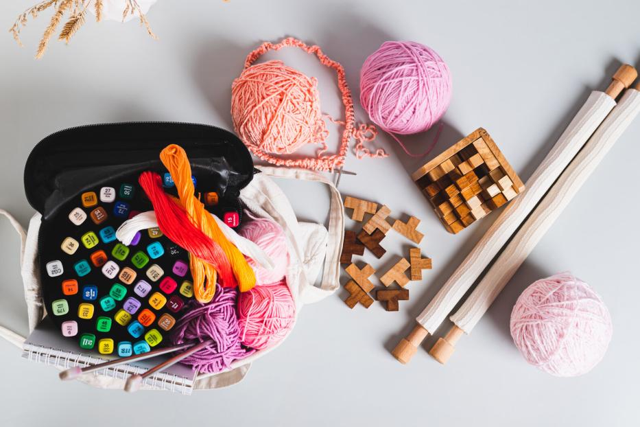Analog bag of yarn, markers, a notebook, knitting needles, and puzzles top view web banner