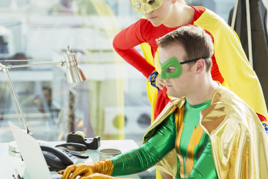 Zwei Erwachsene in Superheldenkostümen arbeiten gemeinsam an einem Schreibtisch mit Laptop in einem modernen Büro.