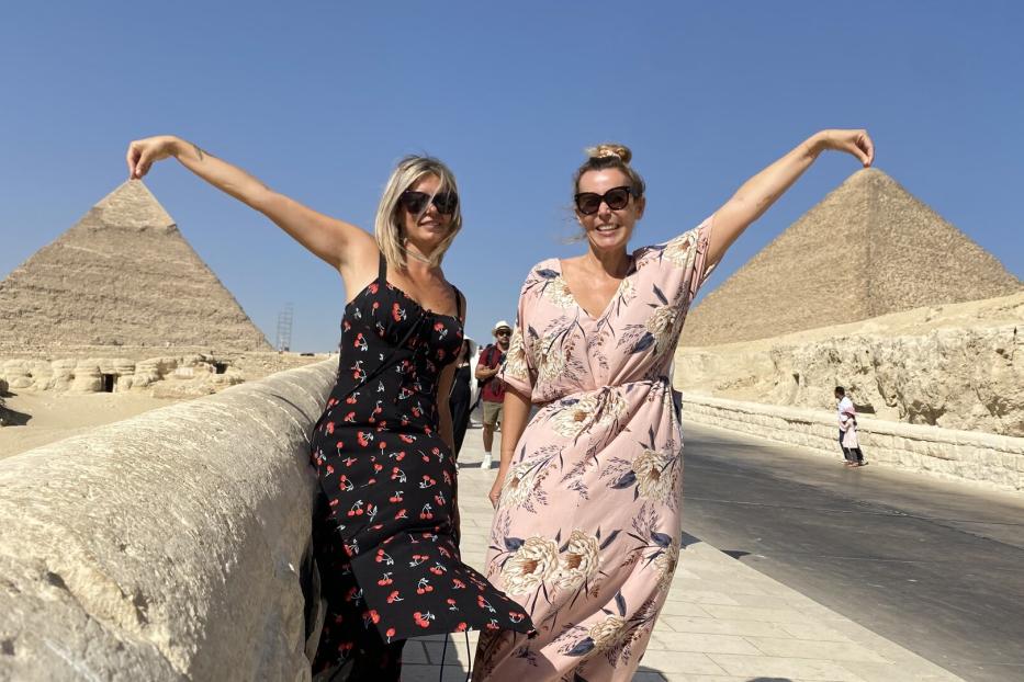 Zwei Frauen posieren vor den Pyramiden von Gizeh mit erhobenen Armen.