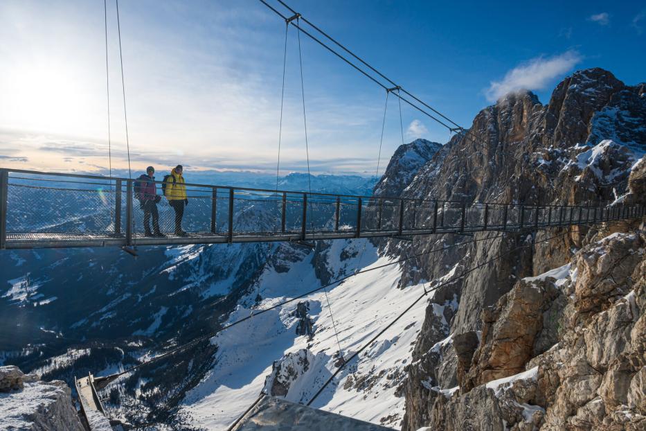 Zwei Personen stehen auf einer Hängebrücke hoch über einer verschneiten Berglandschaft bei Sonnenschein.
