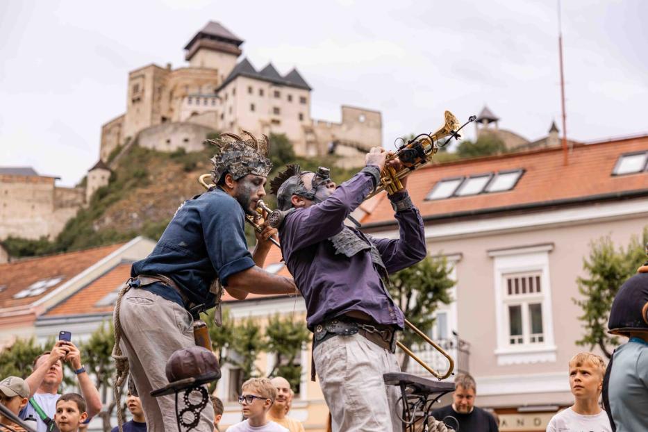 Zwei Musiker mit fantasievollen Kostümen spielen Trompete vor einer Burg und begeistern das Publikum.