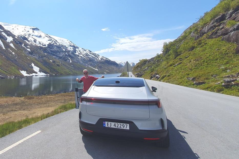 Ein Mann steht neben einem weißen Auto auf einer Straße an einem See, umgeben von Bergen mit Schnee und grüner Vegetation.