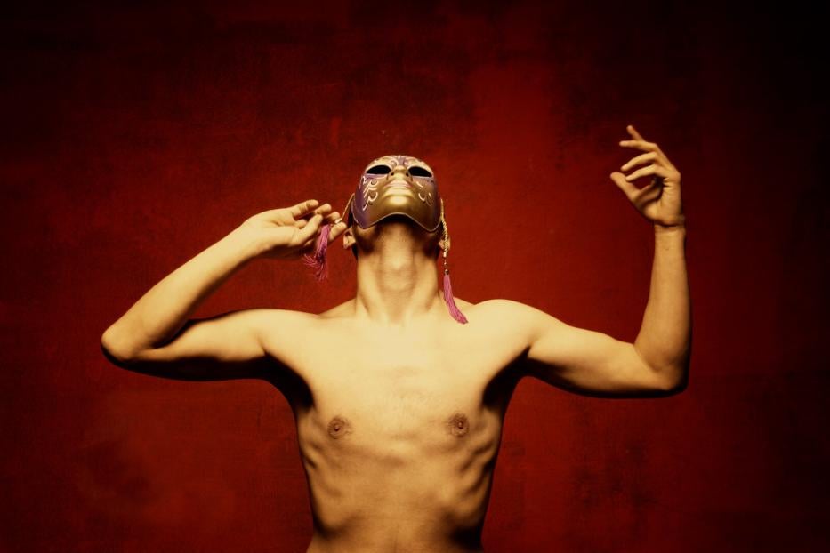 Oberkörper einer Person mit ausgebreiteten Armen und gold-violetter Maske vor rotem Hintergrund.