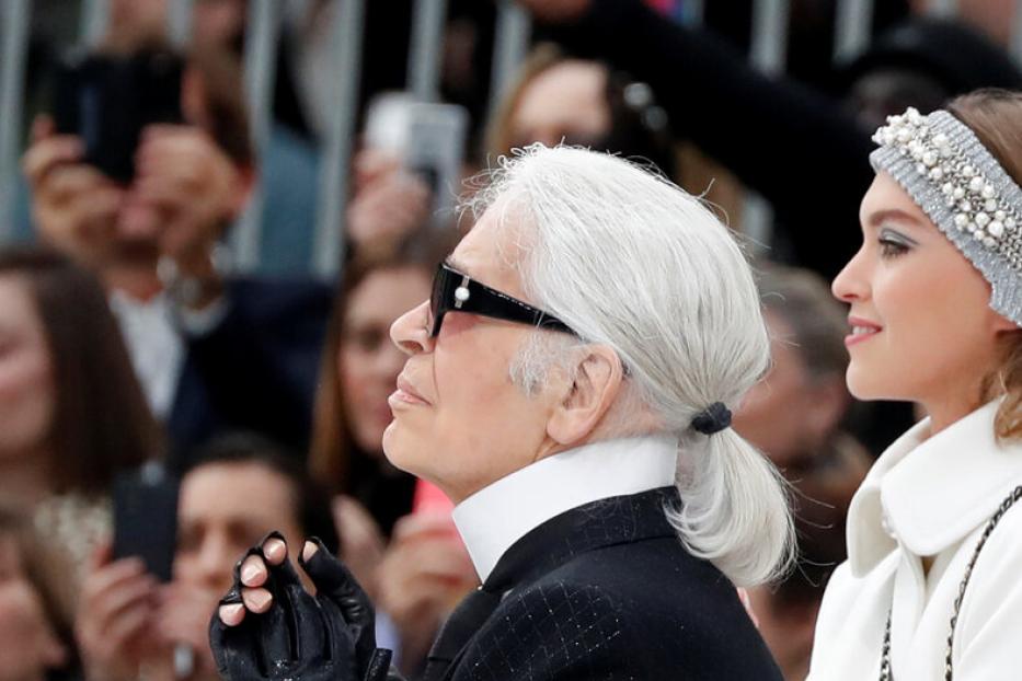Karl Lagerfeld mit Sonnenbrille und Zopf, neben einem Model mit weißem Mantel, blickt aufmerksam in die Ferne.