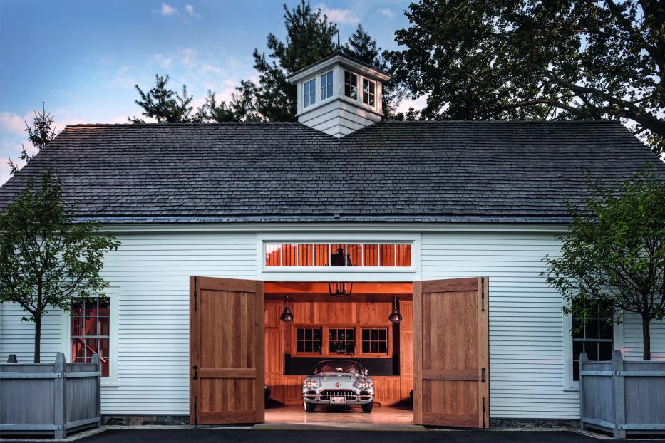 Ein Oldtimer steht in einer offenen, weiß gestrichenen Garage mit Holztoren und gepflegtem Außenbereich.