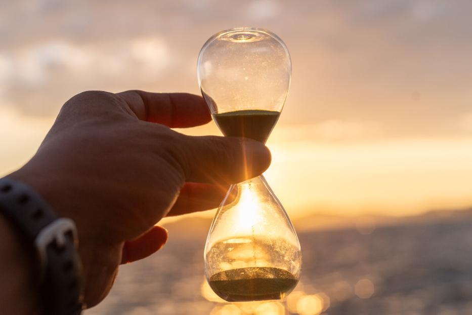Eine Hand hält eine Sanduhr vor einem sonnigen Himmel.